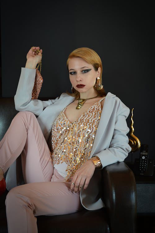 Serious female model in fancy outfit and accessories sitting on sofa with handbag and looking at camera