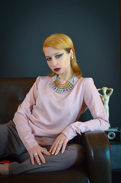 Confident female model wearing blouse and pants with jewelry sitting on leather couch and looking at camera