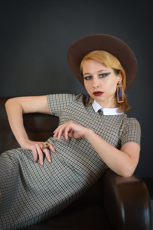 Charismatic female wearing dress and hat with accessories sitting on leather couch and looking at camera