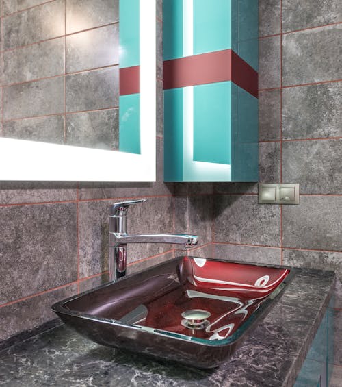 Stylish red sink with faucet on marble counter near tiled wall with illuminated mirror with reflection placed in modern bathroom