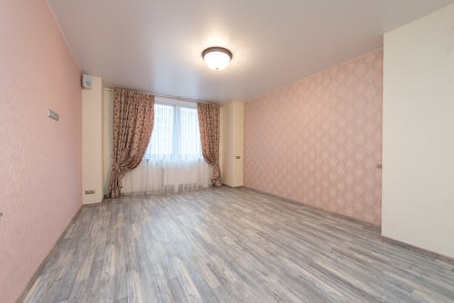 Empty spacious room with laminate and window with colorful curtains between walls with pink ornamental wallpapers and glowing white chandelier