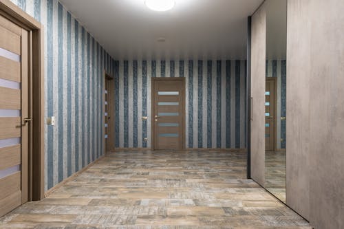 Stylish hallway with mirror on wardrobe and various wooden doors near walls with striped wallpaper in modern flat with glowing chandelier