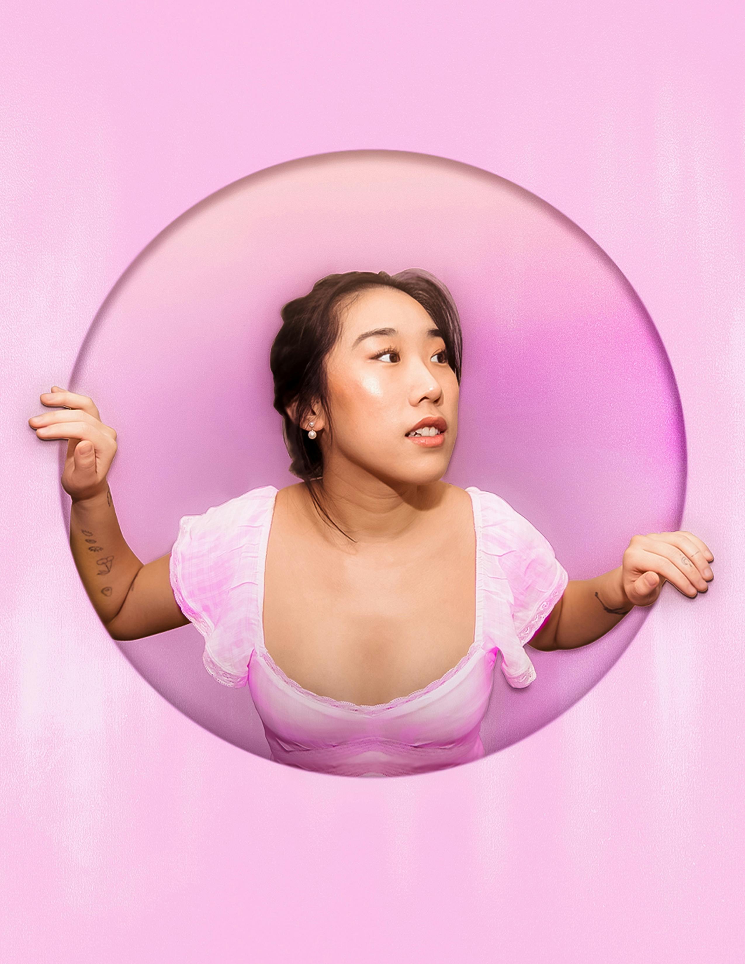 Calm Asian female with dark hair wearing stylish wear looking out of round shaped hole in pink wall in studio