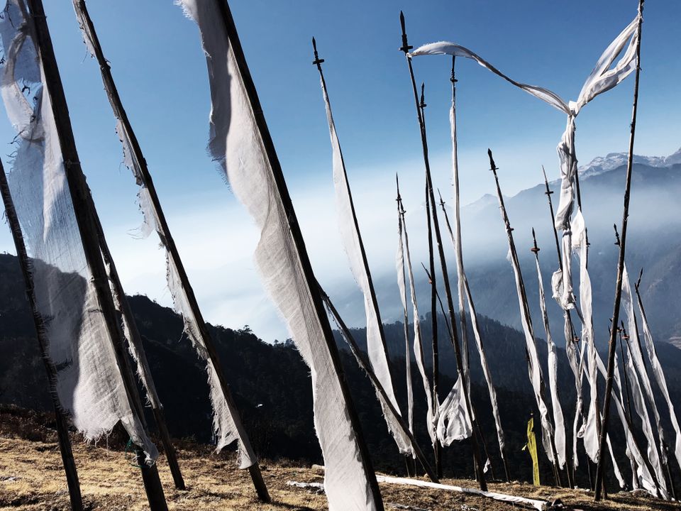 White prayer flags on tall poles along a high ridge above Paro, snow peaks across the haze