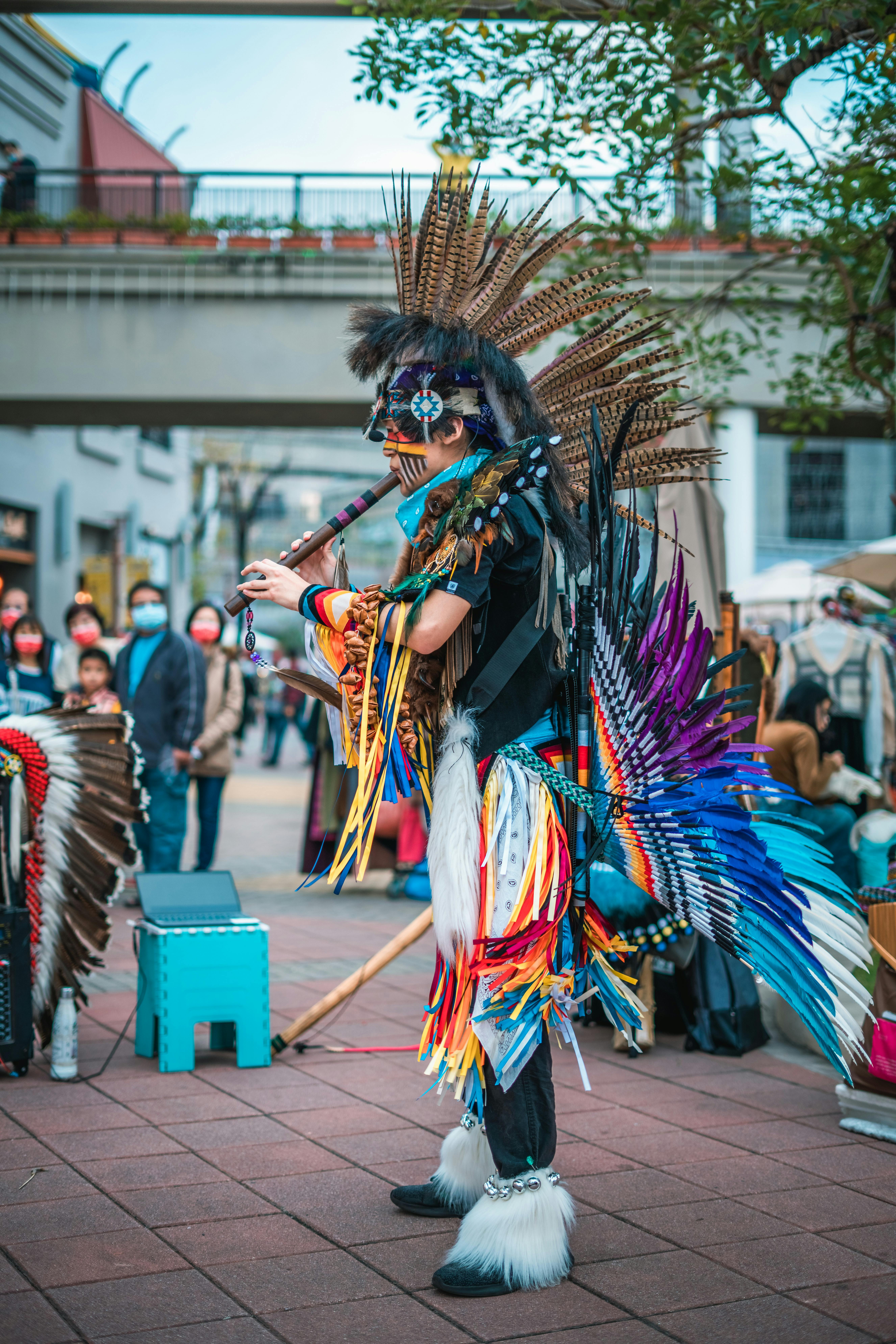 Colorful street performer in feathers and traditional attire playing a flute outdoors.