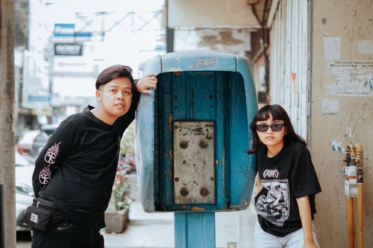 Two young adults posing stylishly by a vintage phone booth in an urban setting.