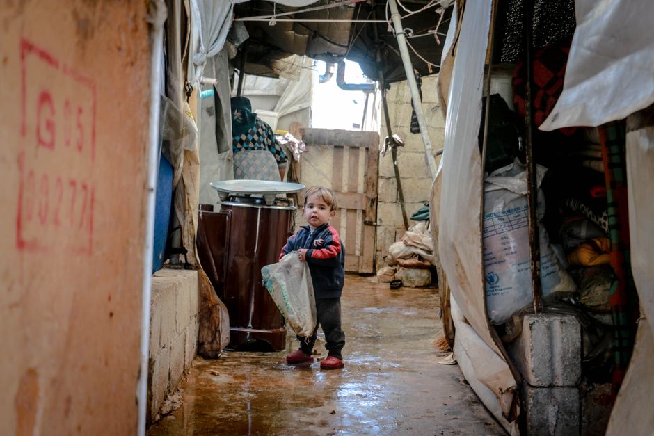 Young child standing in a Syrian refugee camp, holding a plastic bag, indoors.