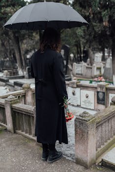 Woman in black coat holding flowers and umbrella at a cemetery, portraying loss and grieving.