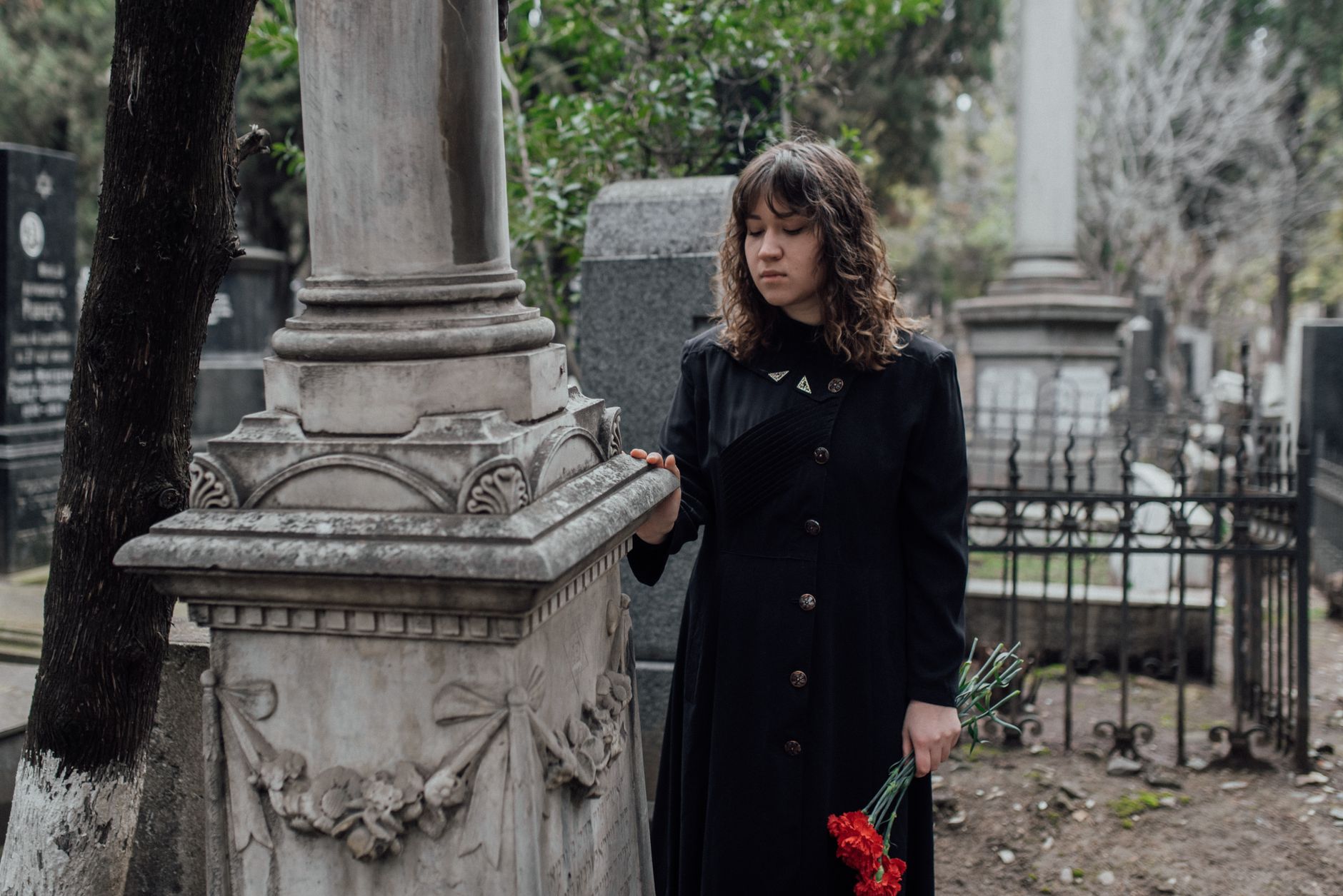 A somber moment as a woman reflects in a cemetery, holding red roses in hand.