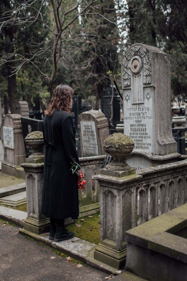 Woman In Black Coat Standing In Front Of A Tombstone With Flowers