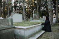 Side view of upset female in black outfit sitting on tiled walkway against tombstone in graveyard