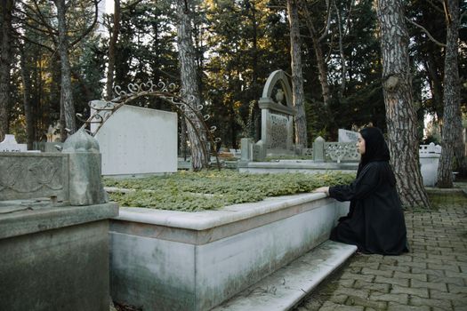 Side view of upset female in black outfit sitting on tiled walkway against tombstone in graveyard