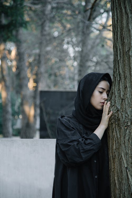 Upset female in black headscarf touching dry tree bark in daylight in cemetery on blurred background