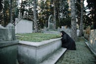 Side view of hopeless female in black wear sitting on tiled pavement against headstone in graveyard