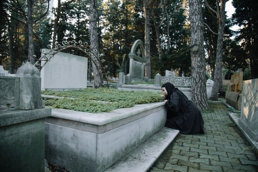 Side view of hopeless female in black wear sitting on tiled pavement against headstone in graveyard