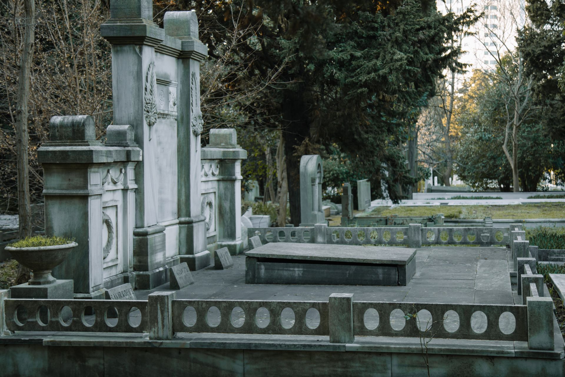 Tombstones with feather decor on fenced grave against overgrown lush green trees in cemetery in daytime