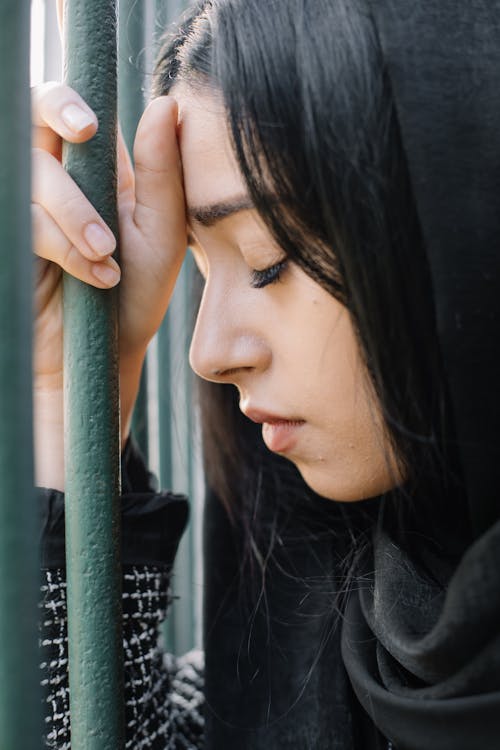 Free Side view of crop young sorrowful female with closed eyes against metal fence in daylight Stock Photo