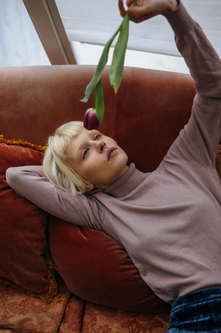 Blonde woman in bohemian attire holding a tulip, reclining on a sofa with a serene expression.