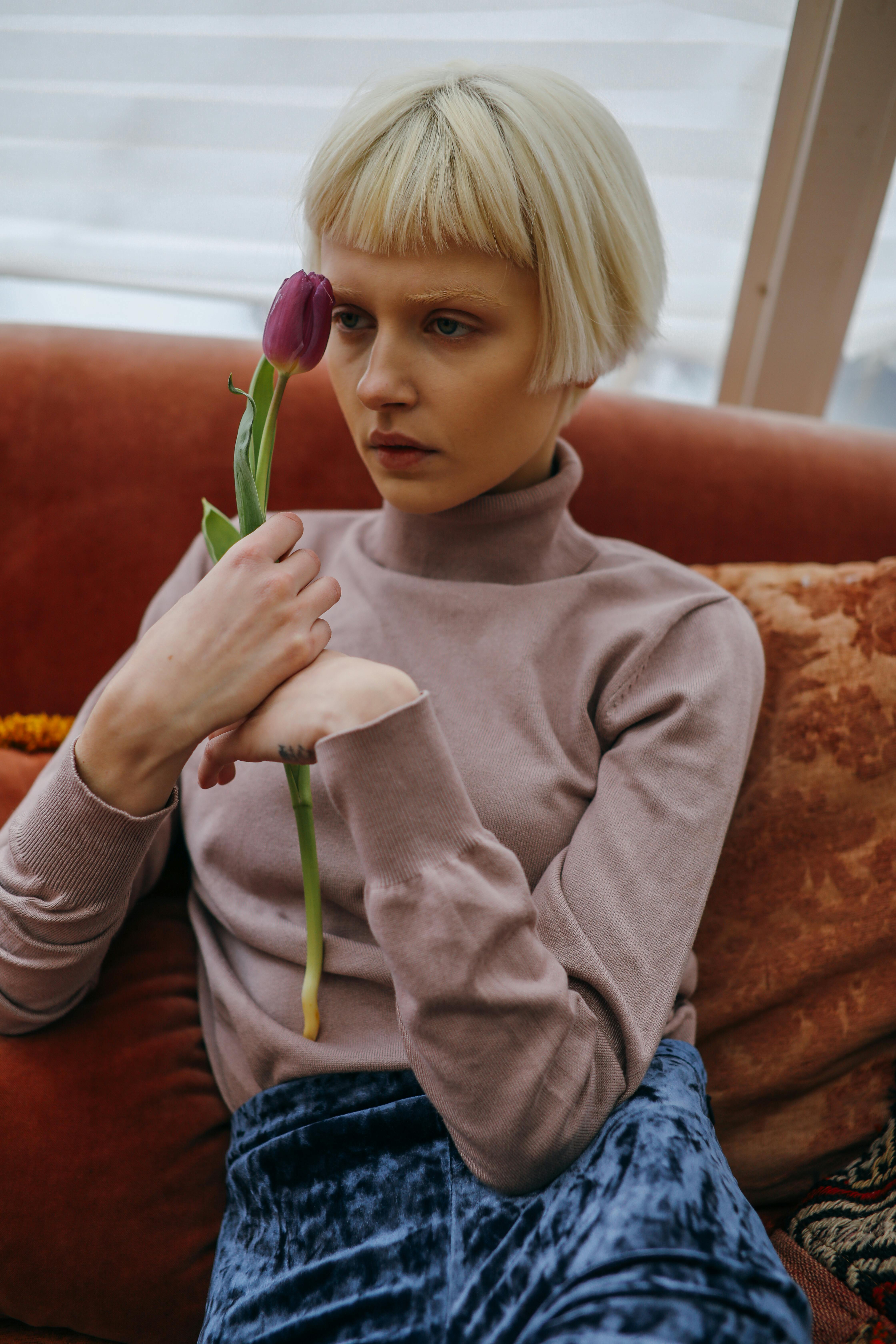 A thoughtful woman with blonde hair holding a purple tulip, sitting on a vintage couch indoors.