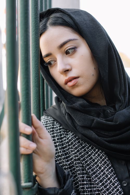 Free Young melancholic female in outerwear with checkered ornament and black headscarf looking away against metal fence outdoors Stock Photo