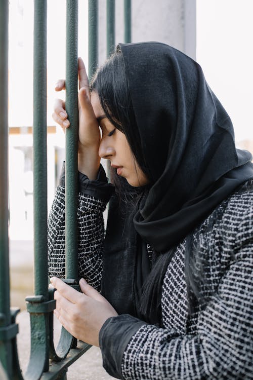 Free Young upset female with closed eyes in outerwear leaning on metal fence in daytime outdoors Stock Photo