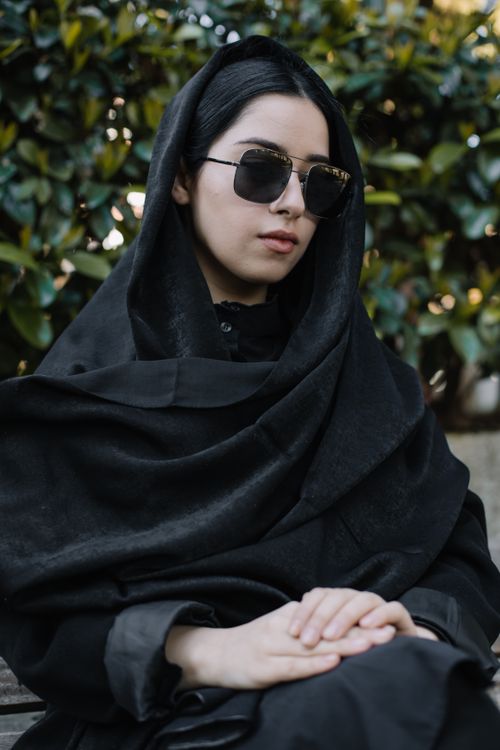 Free Upset female wearing mourning black outfit and sunglasses sitting against bushes with green foliage Stock Photo