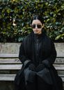 Melancholic female in black outfit and sunglasses sitting on bench and suffering from death of close person