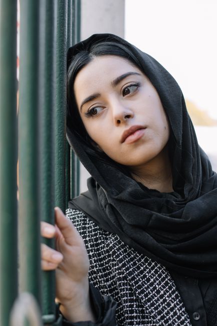 Upset young female in black headscarf leaning head on metal fence and looking away sadly