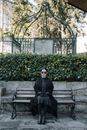 Full body of unemotional female in sunglasses sitting on bench against bushes in cemetery