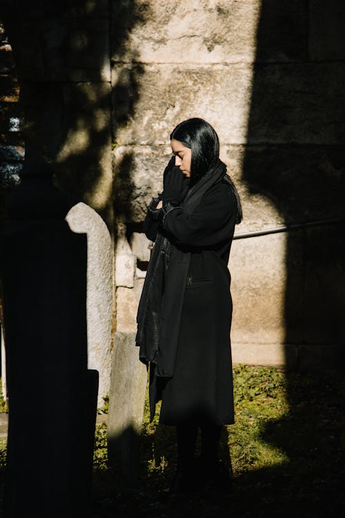 Free Side view full body of female in black mourning clothes standing near grave and praying with closed eyes Stock Photo
