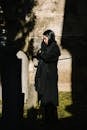 Side view full body of female in black mourning clothes standing near grave and praying with closed eyes