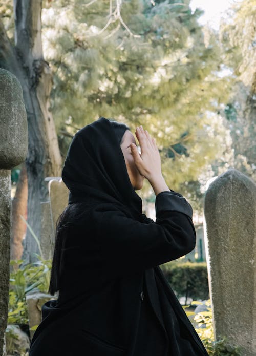 Side view of anonymous female in black clothes with headwear sitting near tombstones with raised hands during traditional ritual on cemetery