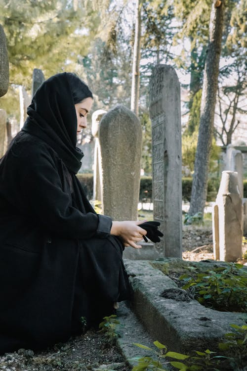 Free Side view full body of calm ethnic female with gloves wearing black clothes and headdress squatting near shabby tombstone on cemetery Stock Photo