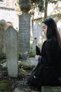 Side view of unhappy ethnic female in black clothes looking down while sitting near stone tombstone on cemetery with abundance of old graves