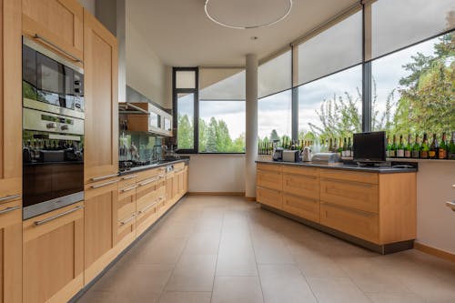 Modern cupboards with contemporary appliances placed near cabinet with kitchenware at big window overlooking green trees in stylish light kitchen