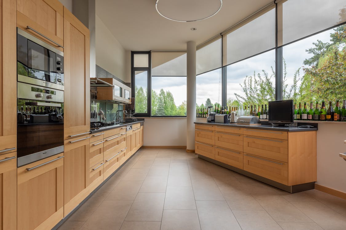 Modern cupboards with contemporary appliances placed near cabinet with kitchenware at big window overlooking green trees in stylish light kitchen