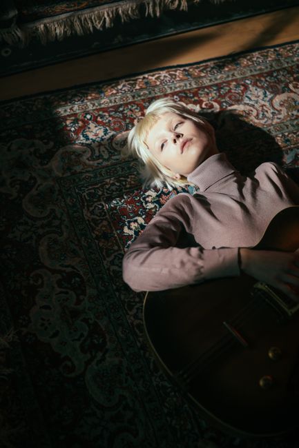 A woman with blond hair lies on a rug holding a guitar, bathed in light and shadow.