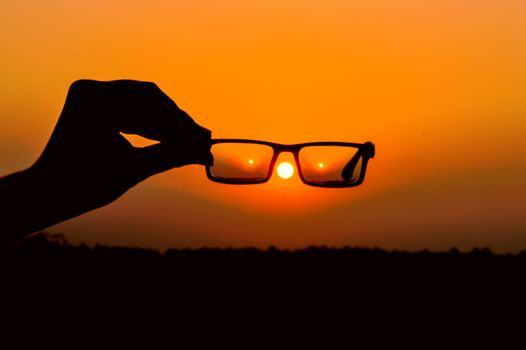 Hand holds eyeglasses against a glowing orange sunset, creating a captivating silhouette effect.