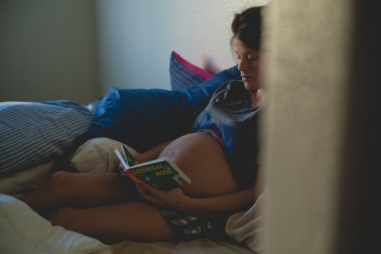 Pregnant Woman Sitting On Bed And Reading Book