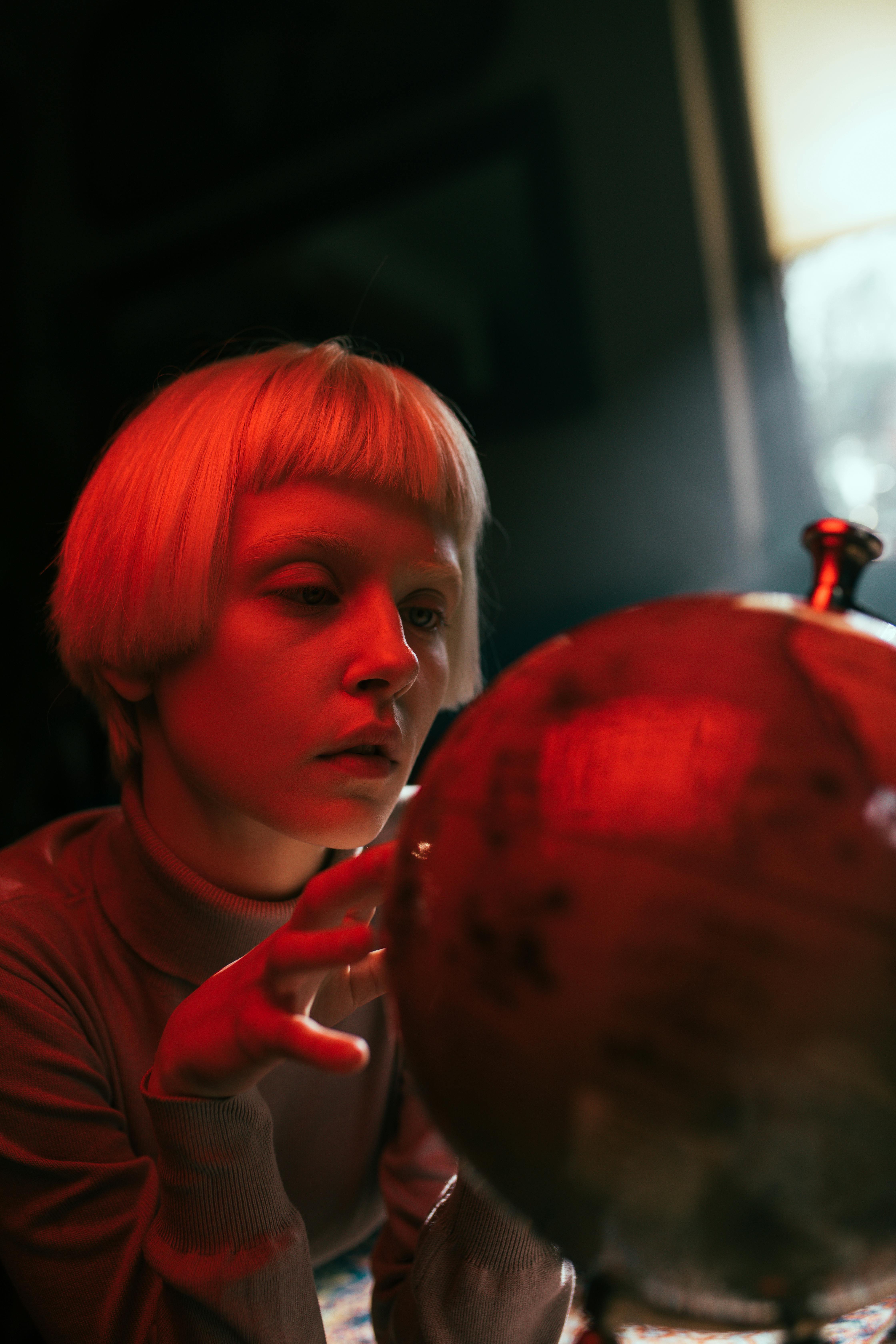 A woman with blonde bangs thoughtfully touches a globe in a dimly lit, cozy room.