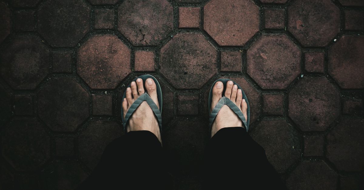 A top-down view of feet wearing flip flops standing on textured red brick pavement.