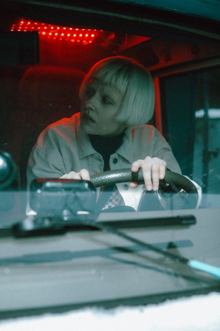 Caucasian woman with blonde hair driving a van illuminated by red interior lights, looking to the side.