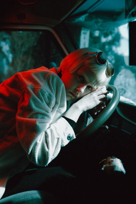 Blonde woman in white jacket artfully poses inside vintage car with colored lighting.