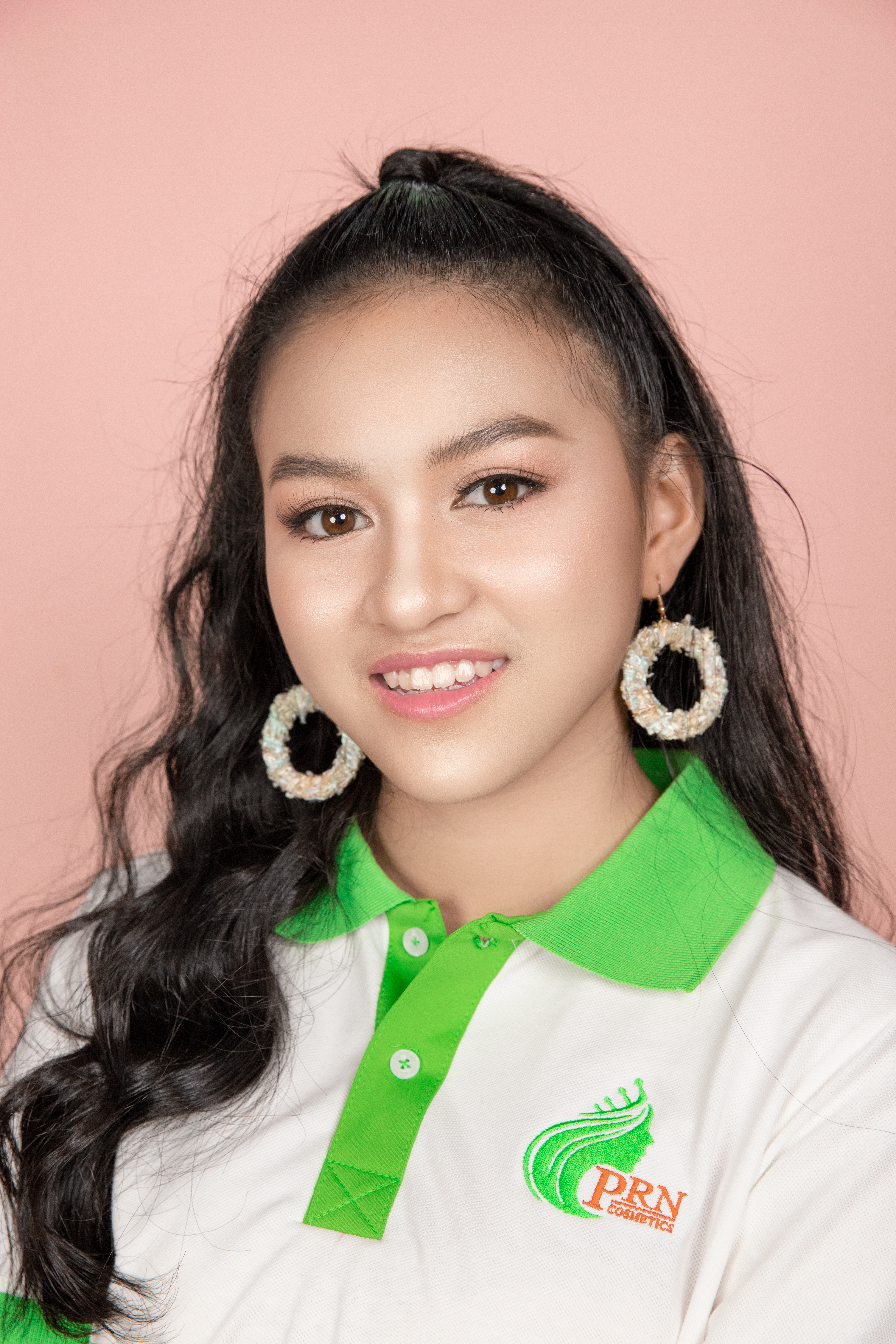 Portrait of a smiling woman with long hair and hoop earrings against a pink background.