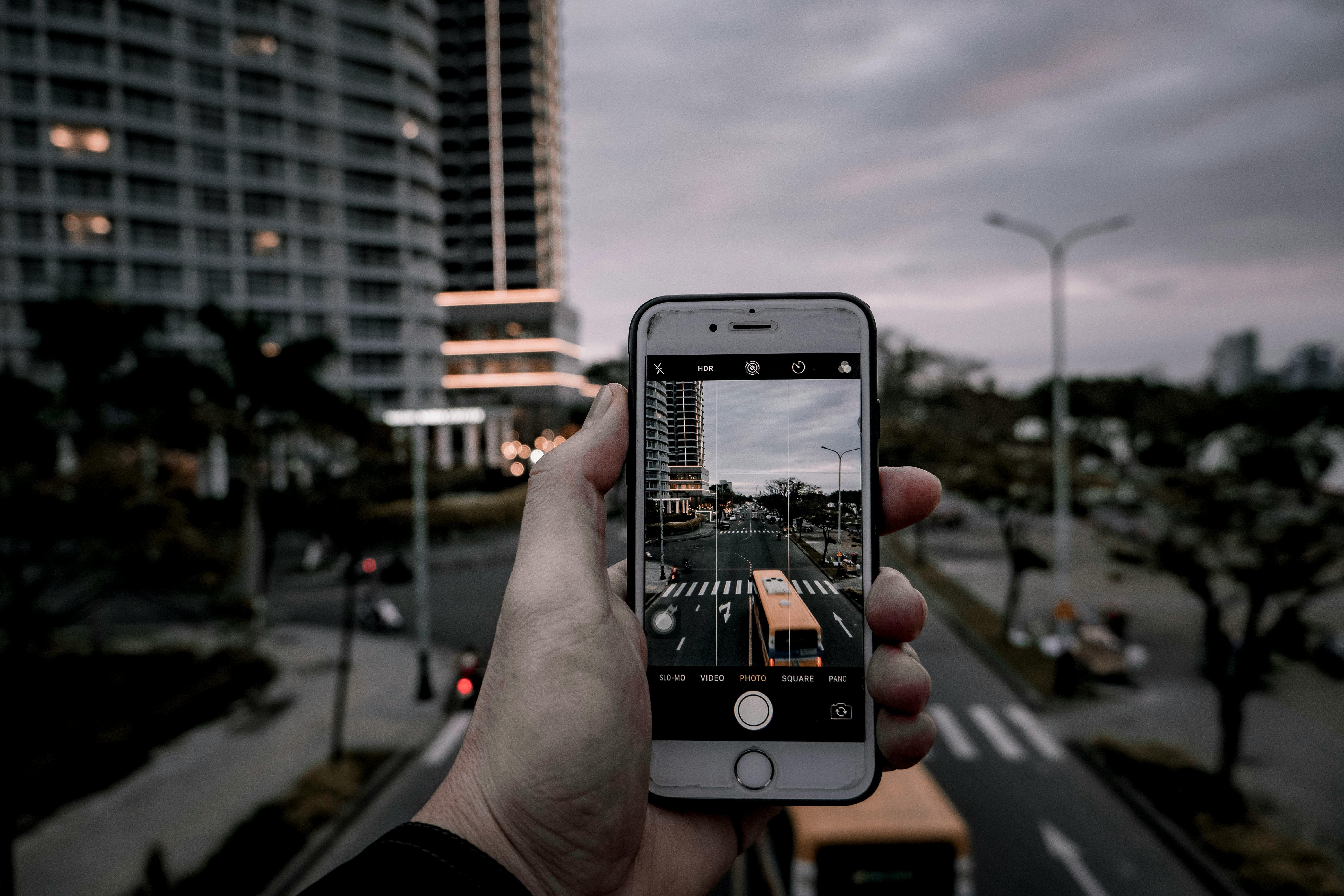 A hand holding a smartphone capturing a city street and buildings in an urban setting.