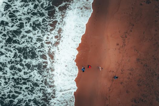 Drone view of foamy waves of stormy ocean rolling on sandy beach near unrecognizable travelers admiring nature