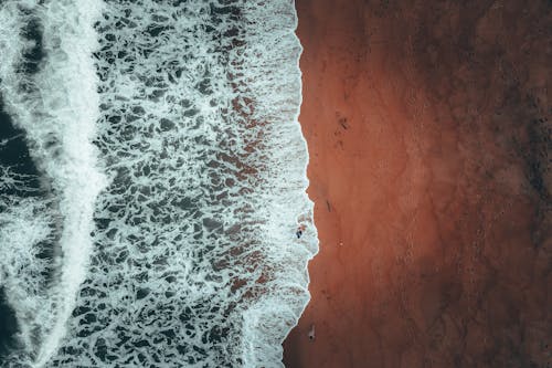 Aerial view of distant unrecognizable tourist standing in foamy water of powerful wavy ocean near sandy beach