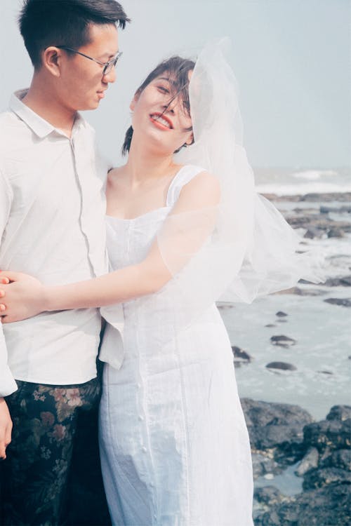 Young couple in casual wedding attire embracing by a rocky seaside, exuding a carefree vibe.