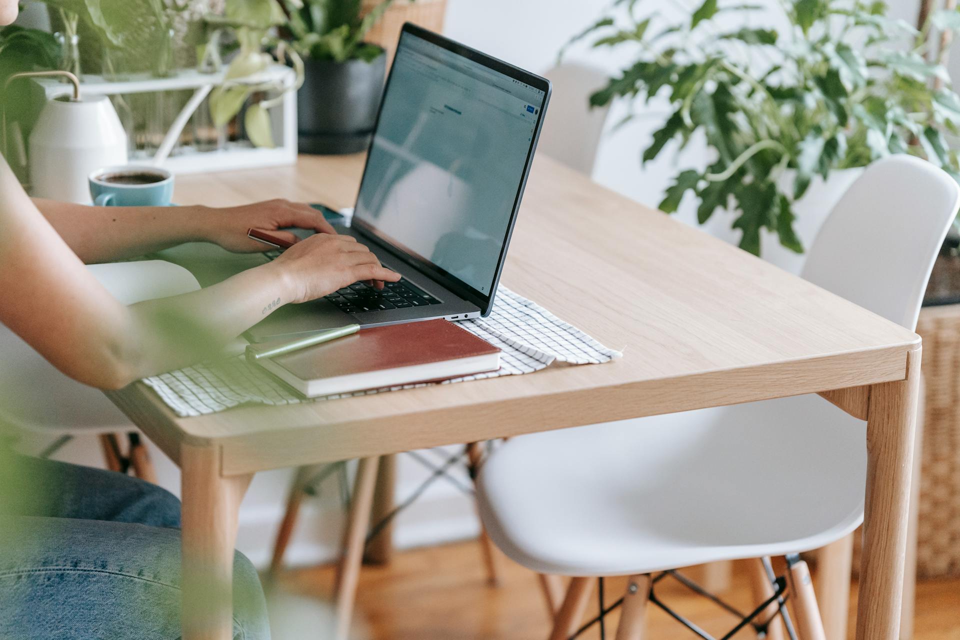 Crop unrecognizable female freelancer browsing internet on netbook while sitting at table in light room with green potted plants at home