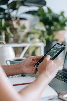 Crop anonymous person surfing cellphone while sitting at table with laptop in light room with green potted plants on blurred background